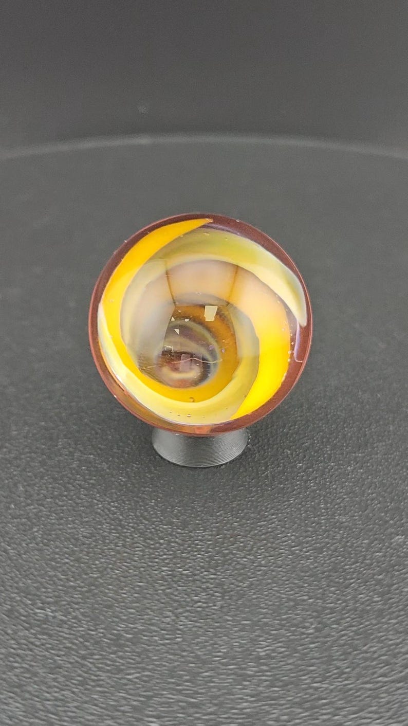 Puede incluir: Una canica de vidrio esf&eacute;rica con un patr&oacute;n interno en espiral de cintas de color amarillo brillante y blanco transl&uacute;cido, encerradas en una capa exterior transparente. La canica se apoya sobre una peque&ntilde;a base circular de color oscuro.