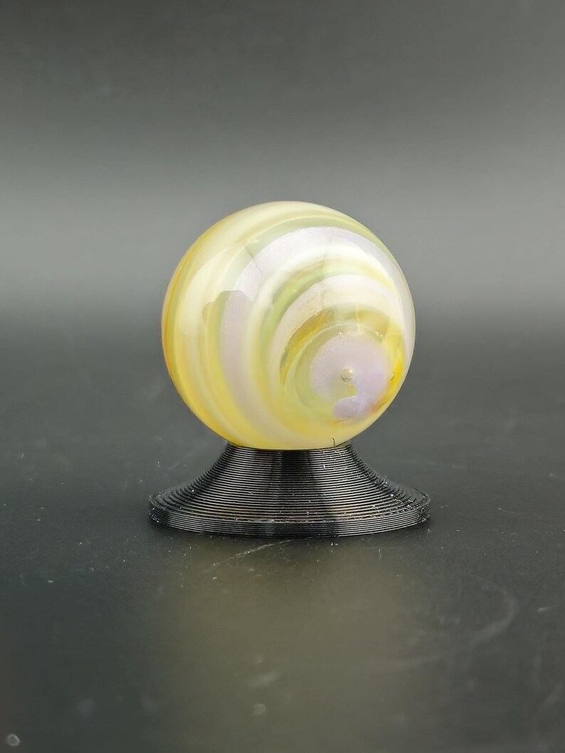 May include: A decorative glass marble with yellow, white, and purple swirls, resting on a black, spiral-shaped stand. The marble has a glossy finish and is a sphere shape.