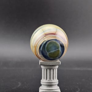 May include: A decorative glass orb with swirling colors of tan, brown, and blue, resting on a small, gray, classical-style pedestal. The orb has a central dark blue and green core.