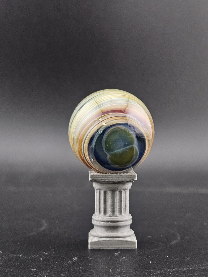 May include: A decorative glass orb with swirling colors of tan, brown, and blue, resting on a small, gray, classical-style pedestal. The orb has a central dark blue and green core.