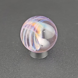 May include: A spherical glass marble featuring a translucent light purple base with swirling internal patterns of white, blue, and orange. The marble is supported by a small, circular, dark gray stand, showcasing a smooth, polished surface.