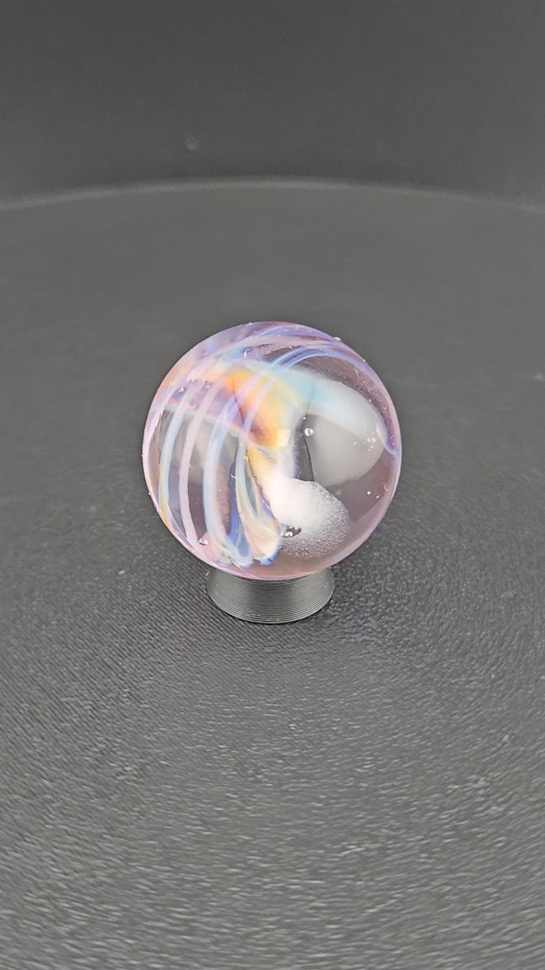 May include: A spherical glass marble featuring a translucent light purple base with swirling internal patterns of white, blue, and orange. The marble is supported by a small, circular, dark gray stand, showcasing a smooth, polished surface.