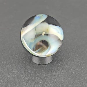 May include: A spherical glass marble featuring a clear outer layer with swirling internal patterns of white, light blue, and dark brown. The marble is resting on a small, circular, dark gray stand that provides a stable base for the object.