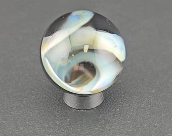May include: A spherical glass marble featuring a clear outer layer with swirling internal patterns of white, light blue, and dark brown. The marble is resting on a small, circular, dark gray stand that provides a stable base for the object.