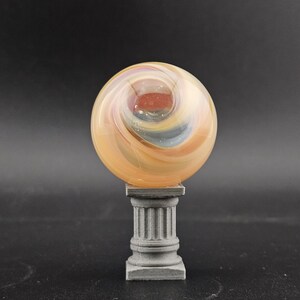 May include: A decorative glass orb with swirling orange, yellow, and blue colors, resting on a gray, column-shaped pedestal. The orb has a central red core and a polished finish, creating a unique display piece.