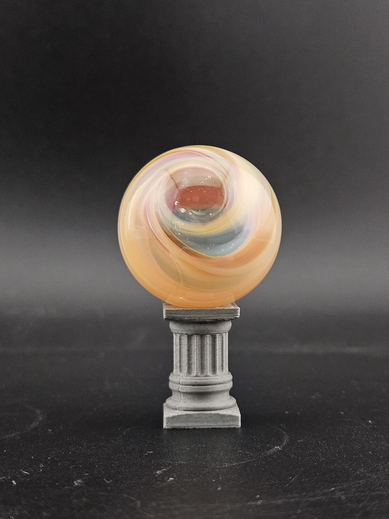 May include: A decorative glass orb with swirling orange, yellow, and blue colors, resting on a gray, column-shaped pedestal. The orb has a central red core and a polished finish, creating a unique display piece.