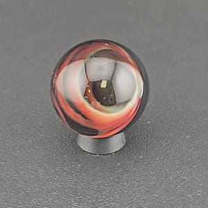 May include: A spherical glass marble featuring swirling patterns of deep black, vibrant orange, and translucent white. The surface is smooth and reflective, showcasing internal layers of color that create a dynamic, multi-dimensional appearance.