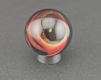 May include: A spherical glass marble featuring swirling patterns of deep black, vibrant orange, and translucent white. The surface is smooth and reflective, showcasing internal layers of color that create a dynamic, multi-dimensional appearance.