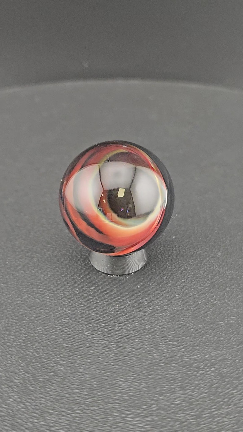 May include: A spherical glass marble featuring swirling patterns of deep black, vibrant orange, and translucent white. The surface is smooth and reflective, showcasing internal layers of colour that create a dynamic, multi-dimensional appearance.