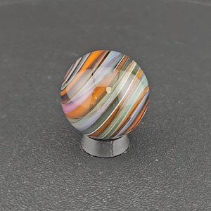 May include: A spherical glass marble featuring a swirling pattern of orange, light purple, dark green, and white stripes. The marble is resting on a small, circular, dark gray stand that provides a stable base for the round object.