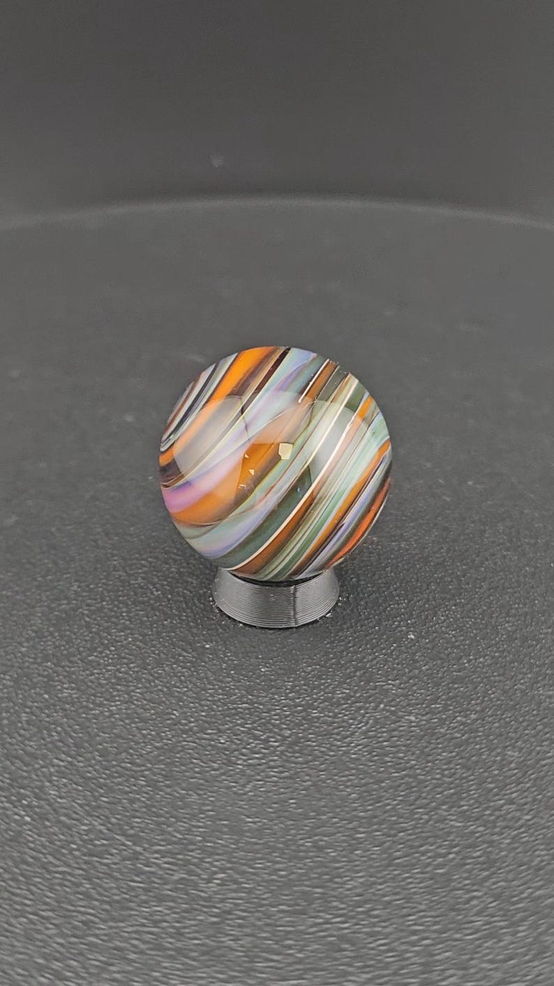 May include: A spherical glass marble featuring a swirling pattern of orange, light purple, dark green, and white stripes. The marble is resting on a small, circular, dark gray stand that provides a stable base for the round object.