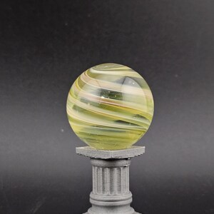 May include: A decorative glass orb with swirling yellow, green, and white colors, resting on a small, gray, classical-style pedestal. The orb is transparent, allowing light to pass through.