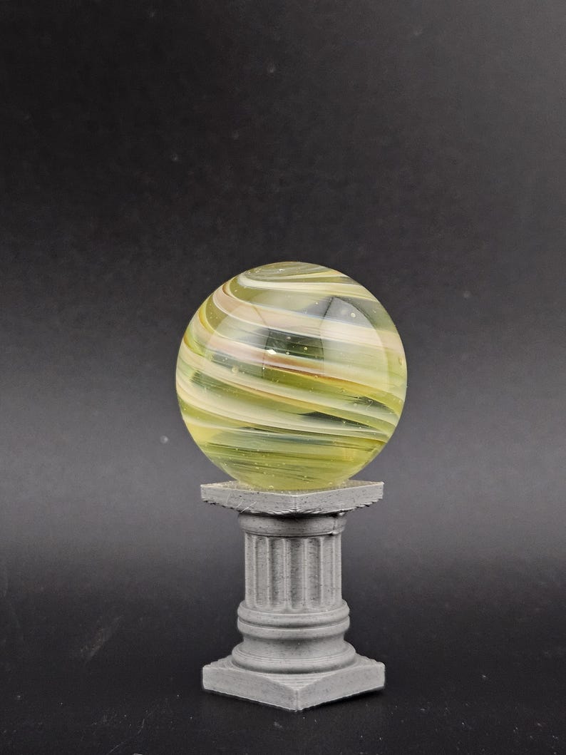 May include: A decorative glass orb with swirling yellow, green, and white colors, resting on a small, gray, classical-style pedestal. The orb is transparent, allowing light to pass through.
