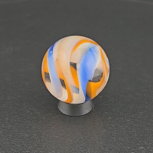 May include: A spherical glass marble featuring a swirling pattern of opaque white, vibrant orange, and bright blue ribbons set against a clear base. The marble is resting on a small, circular black stand, showcasing its smooth, polished surface.
