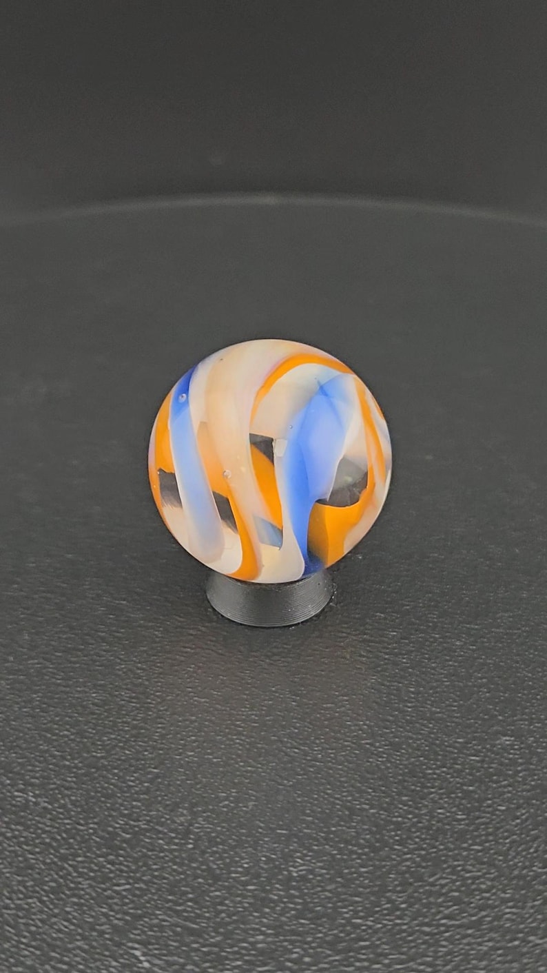 May include: A spherical glass marble featuring a swirling pattern of opaque white, vibrant orange, and bright blue ribbons set against a clear base. The marble is resting on a small, circular black stand, showcasing its smooth, polished surface.