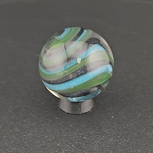 May include: A spherical glass marble featuring swirling bands of translucent light blue, opaque green, and dark charcoal gray. The surface is smooth and reflective, showcasing a complex internal pattern of layered colors that wrap around the orb.