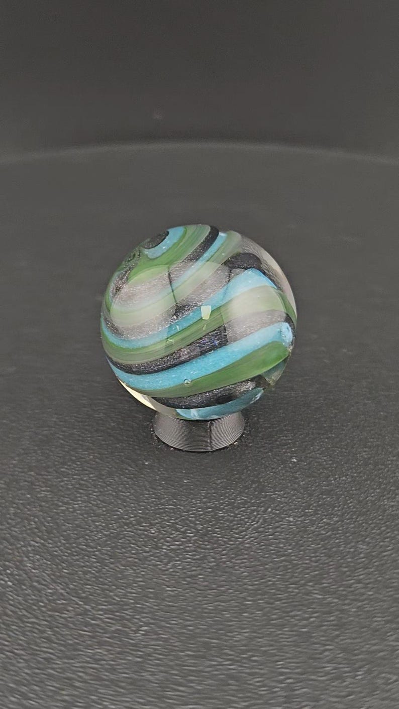 May include: A spherical glass marble featuring swirling bands of translucent light blue, opaque green, and dark charcoal gray. The surface is smooth and reflective, showcasing a complex internal pattern of layered colors that wrap around the orb.