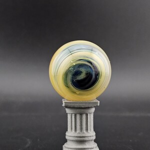 May include: A glass marble with yellow, blue, and white swirls, resting on a small, gray, fluted column. The marble has a glossy finish and a dark blue center. The column is a miniature replica of a classical architectural element.