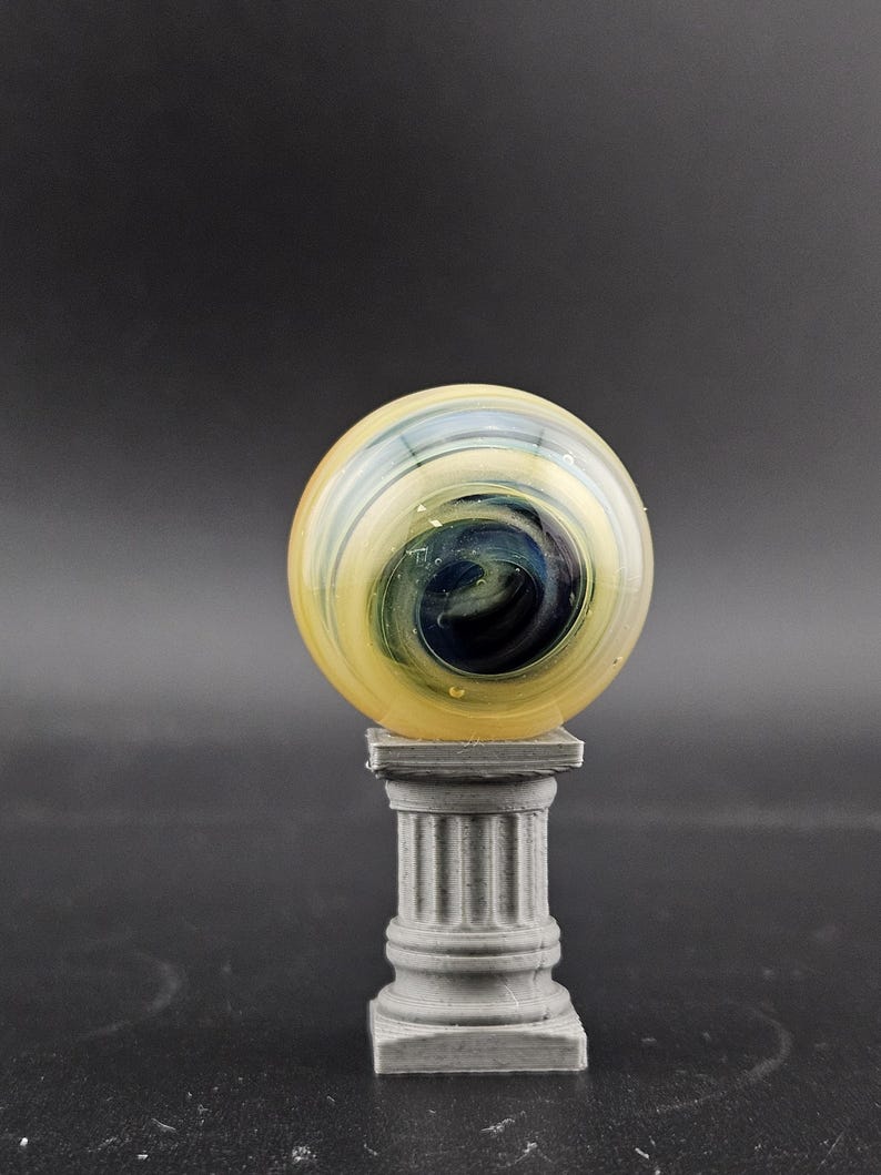 May include: A glass marble with yellow, blue, and white swirls, resting on a small, gray, fluted column. The marble has a glossy finish and a dark blue center. The column is a miniature replica of a classical architectural element.
