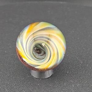 May include: A spherical glass marble featuring a swirling vortex pattern of yellow, white, and translucent blue colors. The center of the sphere displays a deep dark brown core, while the base of the marble is accented with a textured reddish-brown surface.
