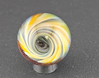 May include: A spherical glass marble featuring a swirling vortex pattern of yellow, white, and translucent blue colors. The center of the sphere displays a deep dark brown core, while the base of the marble is accented with a textured reddish-brown surface.