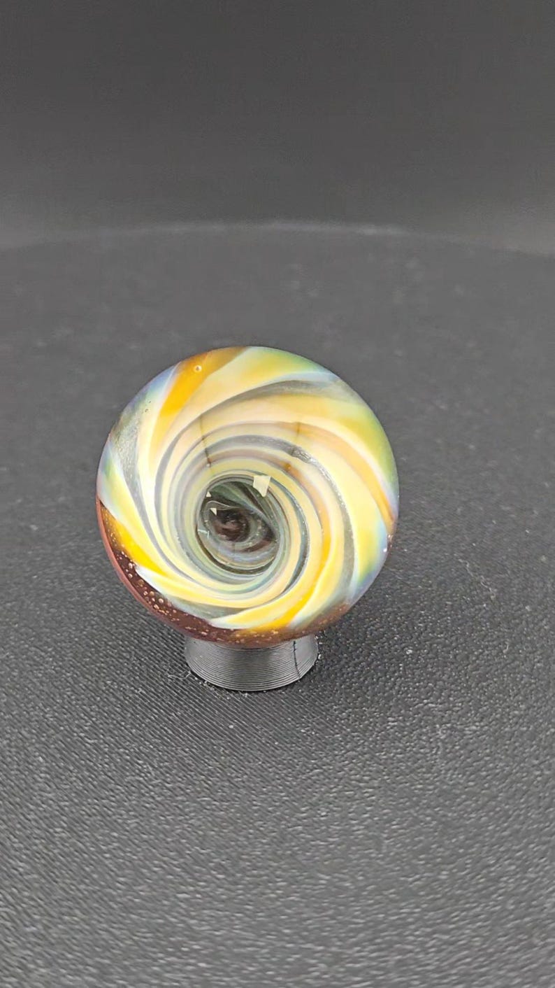 May include: A spherical glass marble featuring a swirling vortex pattern of yellow, white, and translucent blue colors. The center of the sphere displays a deep dark brown core, while the base of the marble is accented with a textured reddish-brown surface.