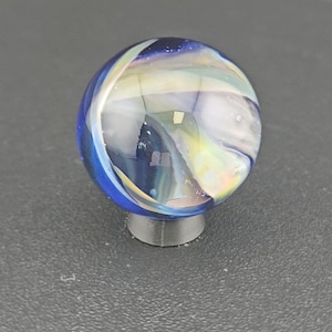 May include: A spherical glass marble featuring a deep blue base color with swirling translucent white and iridescent ribbons throughout the surface. The marble is resting on a small, circular, dark gray stand that supports its rounded shape.