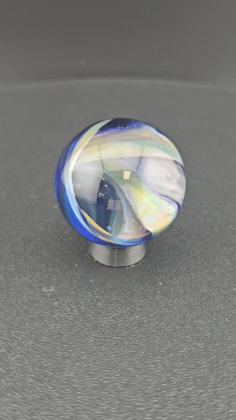May include: A spherical glass marble featuring a deep blue base color with swirling translucent white and iridescent ribbons throughout the surface. The marble is resting on a small, circular, dark gray stand that supports its rounded shape.