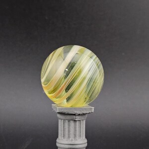 May include: A clear glass marble with yellow and white swirling patterns, resting on a small, gray, classical-style pedestal. The marble is spherical, and the pedestal has a square top and base with a fluted column.