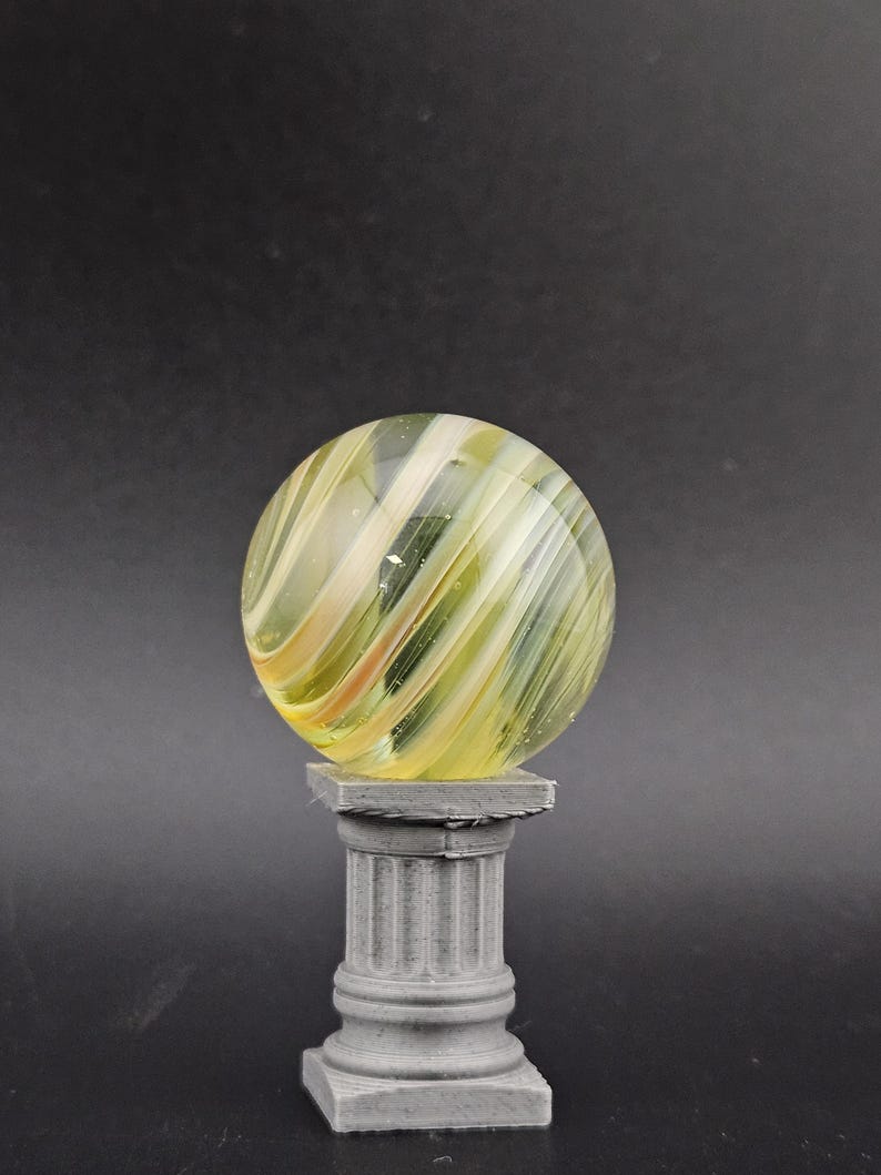 May include: A clear glass marble with yellow and white swirling patterns, resting on a small, gray, classical-style pedestal. The marble is spherical, and the pedestal has a square top and base with a fluted column.