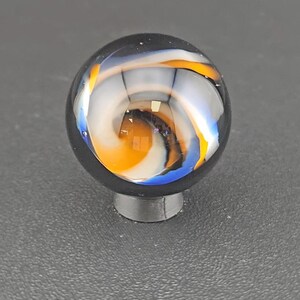 May include: A spherical glass marble featuring a clear outer layer with internal swirling patterns of opaque white, vibrant orange, and deep blue colours. The marble is resting on a small, circular, dark grey stand that supports its round shape.