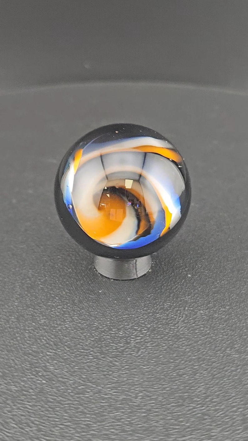 May include: A spherical glass marble featuring a clear outer layer with internal swirling patterns of opaque white, vibrant orange, and deep blue colours. The marble is resting on a small, circular, dark grey stand that supports its round shape.