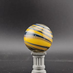 May include: A decorative glass orb with yellow, blue, and black swirling patterns, resting on a small, gray, classical-style pedestal. The orb is spherical, and the pedestal has a fluted column design.