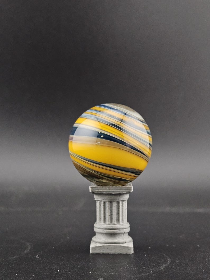 May include: A decorative glass orb with yellow, blue, and black swirling patterns, resting on a small, gray, classical-style pedestal. The orb is spherical, and the pedestal has a fluted column design.