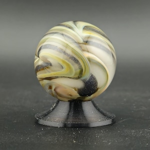 May include: A decorative glass marble with a swirl pattern of white, yellow, and black. The marble sits on a black, wire-like pedestal. The design is artistic and eye-catching, suitable for display. The base is made of wire.