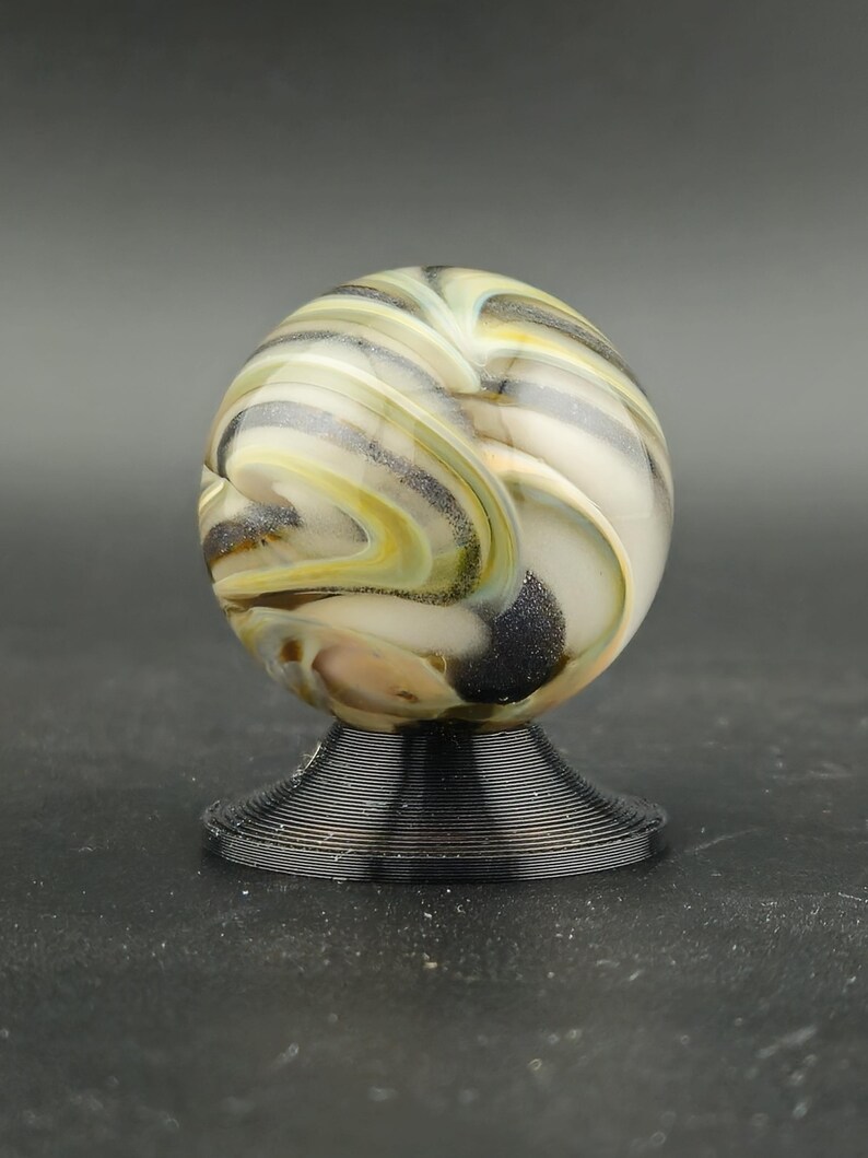 May include: A decorative glass marble with a swirl pattern of white, yellow, and black. The marble sits on a black, wire-like pedestal. The design is artistic and eye-catching, suitable for display. The base is made of wire.