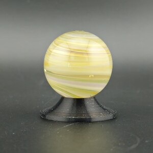 May include: A decorative glass marble with yellow, white, and green swirls, resting on a black, spiral-shaped base. The marble is spherical and appears to be made of translucent glass. The base is made of a dark, possibly metal material.
