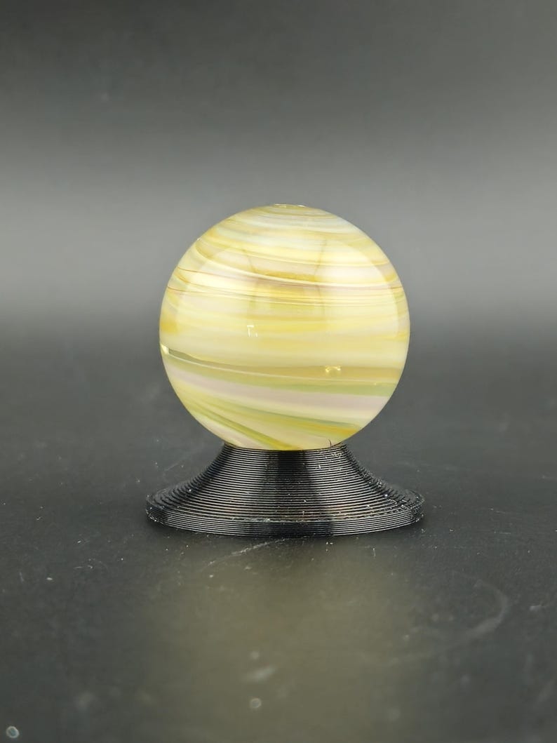 May include: A decorative glass marble with yellow, white, and green swirls, resting on a black, spiral-shaped base. The marble is spherical and appears to be made of translucent glass. The base is made of a dark, possibly metal material.