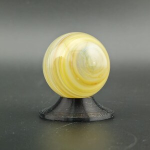 May include: A decorative glass sphere with yellow, white, and blue swirls, resting on a black, spiral-shaped base. The sphere has a glossy finish and is a decorative item.