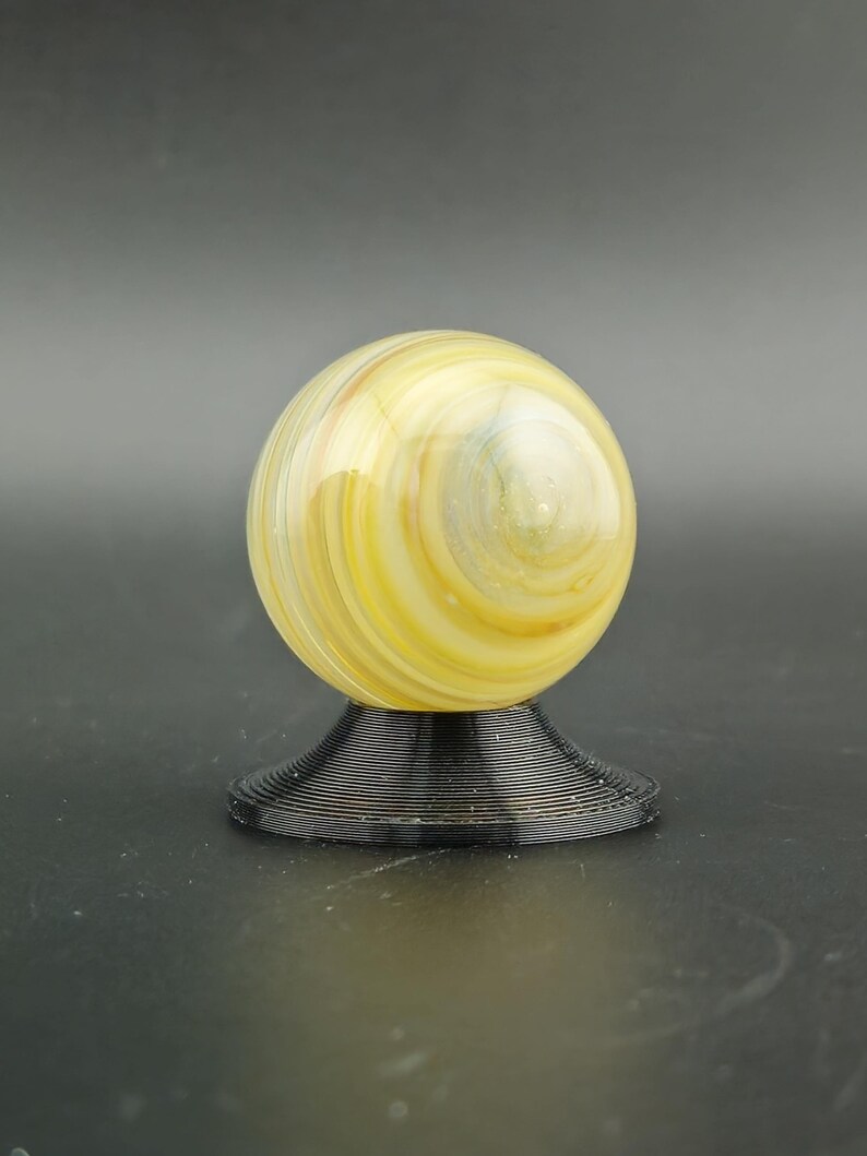 May include: A decorative glass sphere with yellow, white, and blue swirls, resting on a black, spiral-shaped base. The sphere has a glossy finish and is a decorative item.