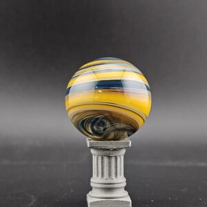May include: A decorative glass marble with yellow, blue, and brown swirling patterns. The marble is displayed on a small, gray, classical-style pedestal. The background is a solid, dark gray.