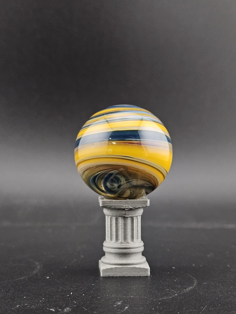 May include: A decorative glass marble with yellow, blue, and brown swirling patterns. The marble is displayed on a small, gray, classical-style pedestal. The background is a solid, dark gray.