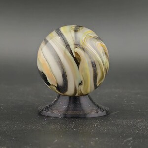 May include: A decorative glass marble with a swirl pattern of cream, yellow, black, and grey. The marble sits on a black, spiral-shaped stand. The background is a solid grey. A unique display piece.