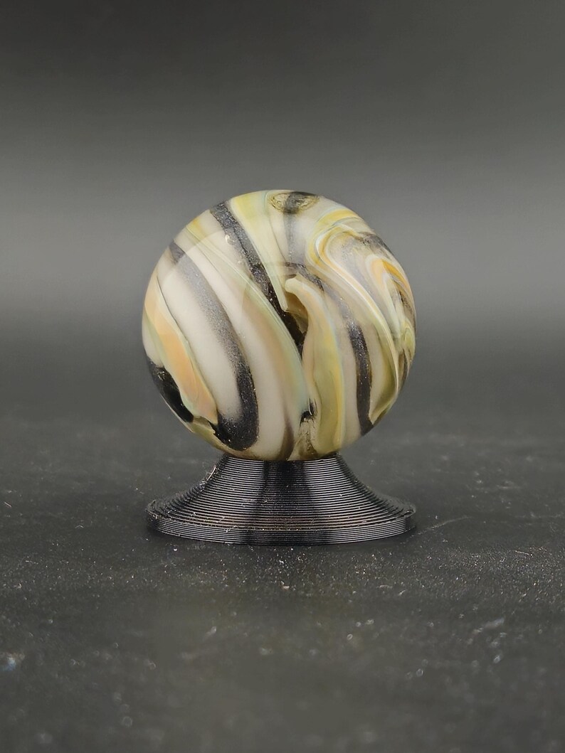 May include: A decorative glass marble with a swirl pattern of cream, yellow, black, and grey. The marble sits on a black, spiral-shaped stand. The background is a solid grey. A unique display piece.