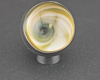 May include: A spherical cabinet knob featuring a swirling pattern of white, yellow, and dark gray glass. The knob is set into a dark metallic base, creating a smooth, rounded profile with a polished finish that reflects light across its surface.