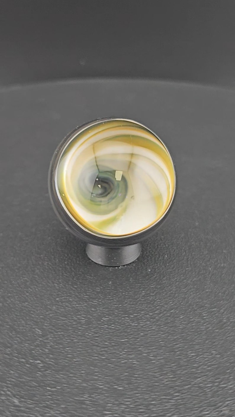 May include: A spherical cabinet knob featuring a swirling pattern of white, yellow, and dark gray glass. The knob is set into a dark metallic base, creating a smooth, rounded profile with a polished finish that reflects light across its surface.