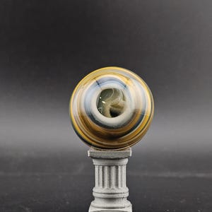 May include: A decorative glass sphere with swirling patterns of gold, blue, and white, resting on a small, gray, fluted pedestal. The sphere has a central opening and is set against a dark background.