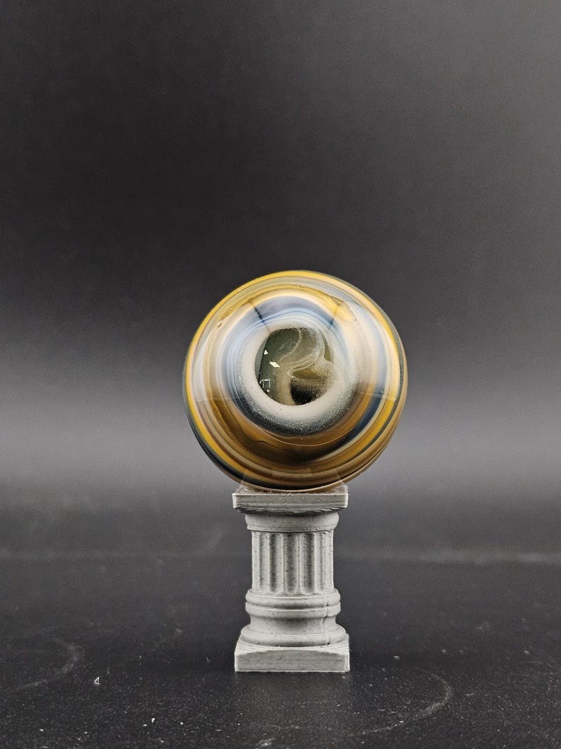 May include: A decorative glass sphere with swirling patterns of gold, blue, and white, resting on a small, gray, fluted pedestal. The sphere has a central opening and is set against a dark background.
