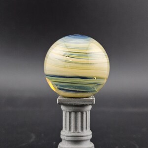 May include: A decorative glass sphere with yellow, blue, and green swirls, resting on a gray, fluted column. The sphere has a polished, reflective surface. The column is a miniature replica of a classical architectural element.