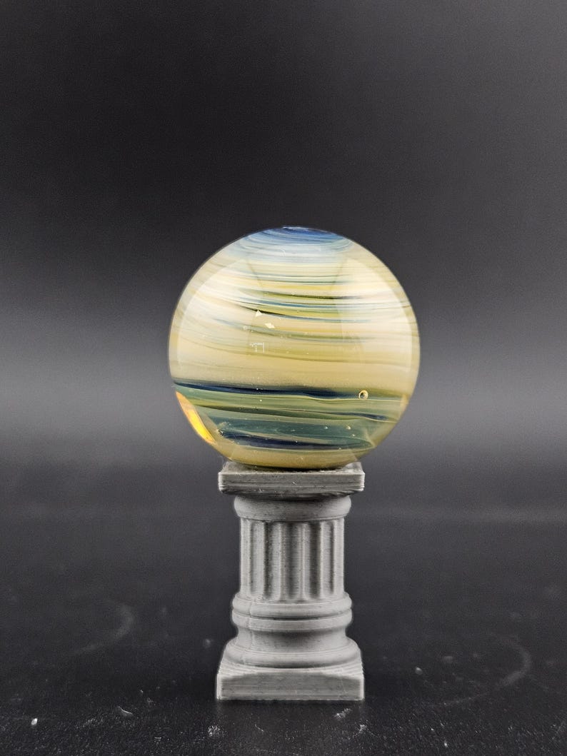 May include: A decorative glass sphere with yellow, blue, and green swirls, resting on a gray, fluted column. The sphere has a polished, reflective surface. The column is a miniature replica of a classical architectural element.